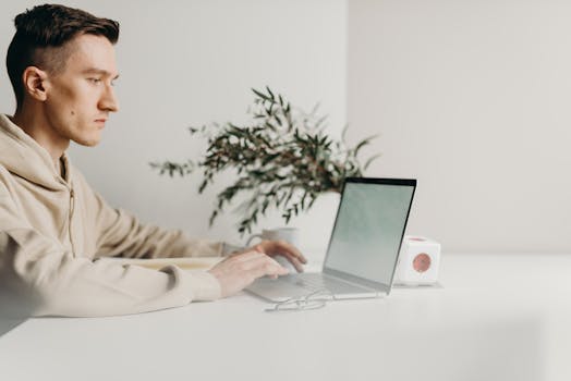 A young man focuses intently on his laptop in a minimalist home office setting.