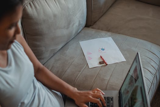 Woman working remotely on a laptop from home with creative sketches nearby, depicting a modern lifestyle.